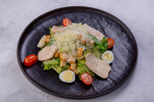 Caesar salad with chicken breast on a dark plate. Home-made chicken fillet, cherry tomatoes, Parmesan cheese, quail eggs, crackers and sauce. European cuisine. Dish from a restaurant or cafe menu. - Foto, Imagen