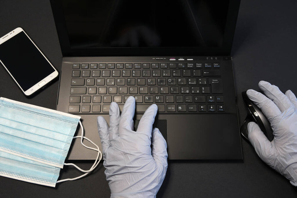 man working on laptop and wearing latex gloves for virus prevention. Man working from home using smartphone and laptop. Surgical face blue masks for coronavirus, covid-19 protection. - Photo, Image