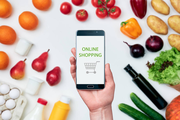 Female hand holding a smartphone next to groceries that surround it: pumpkin, potatoes, onions, cucumbers, lettuce, bell peppers, tomatoes, olive oil, milk, mustard, eggs, yoghurt, oranges, pears - Zdjęcie, obraz