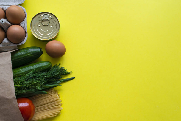 Healthy food delivery during coronavirus. Organic daily meal on yellow background with copy space. Eggs, vegetables, cucumbers, tomatoes, herbs, pasta, oil, canned food in paper bag package, top view. - Fotografie, Obrázek