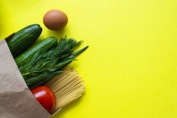 Healthy food delivery during coronavirus. Organic daily meal on yellow background with copy space. Eggs, vegetables, cucumbers, tomatoes, herbs, pasta, oil, canned food in paper bag package, top view. - Photo, Image