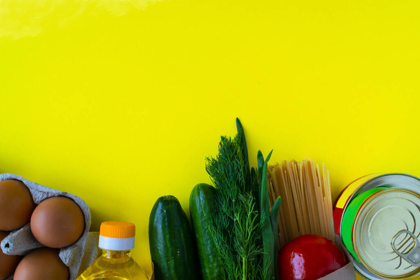 Healthy food delivery during coronavirus. Organic daily meal on yellow background with copy space. Eggs, vegetables, cucumbers, tomatoes, herbs, pasta, oil, canned food in paper bag package, top view. - Photo, Image
