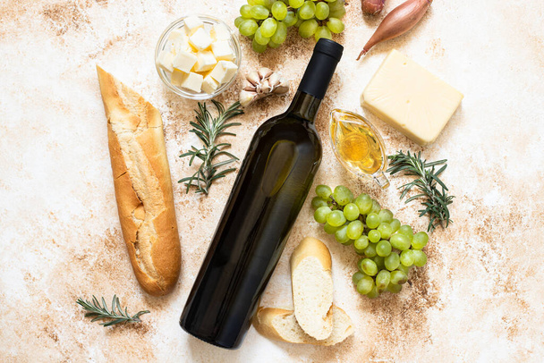 A bottle of white wine, grapes, cheese, butter, honey, baguette, shallot and rosemary. White wine and french cuisine concept. Flat lay, copy space, light marble background - Фото, изображение
