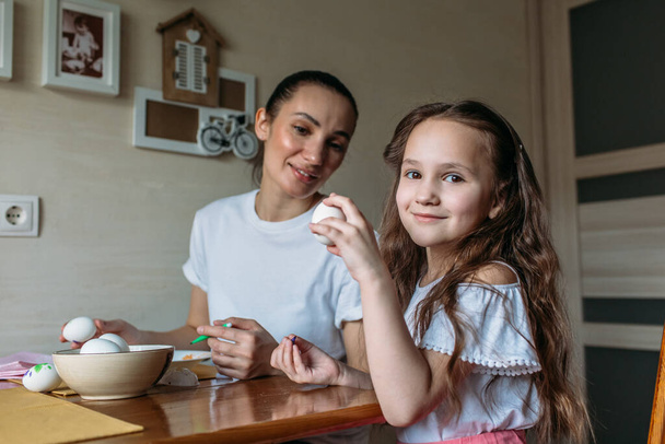 Mom and daughter are sitting in the morning at the kitchen at the kitchen table, on the table is a cup with white eggs, mom and daughter are drawing on white eggs - Foto, Imagen