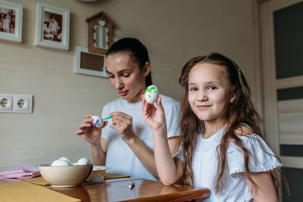 Mom and daughter are sitting in the morning at the kitchen at the kitchen table, on the table is a cup with white eggs, mom and daughter are drawing on white eggs - Foto, Imagen