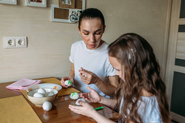 Äiti ja tytär istuvat aamulla keittiössä keittiön pöydällä, pöydällä on kuppi, jossa on valkoisia munia, äiti ja tytär piirtävät valkoisia munia.
 - Valokuva, kuva