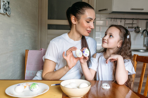 Anya és lánya ülnek reggel a konyhában a konyhaasztalnál, az asztalon van egy csésze fehér tojással, anya és lánya fehér tojást rajzolnak. - Fotó, kép