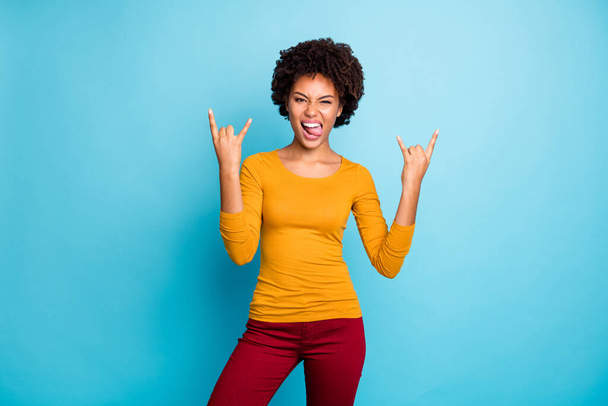 Portrait of cool crazy funky teenage afro american girl make horned symbol enjoy heavy metal concert tongue out grimace face wear stylish jumper red pants isolated over blue color background - Foto, afbeelding