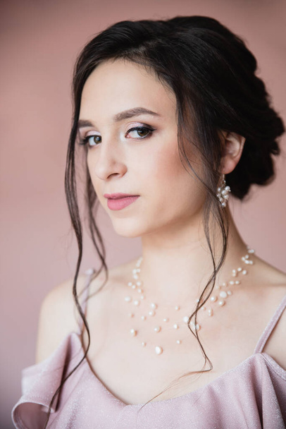 Beautiful young teen girl with perfect make up, styled dark hair, earings and neklace in pink dress on pink background getting ready to prom - 写真・画像