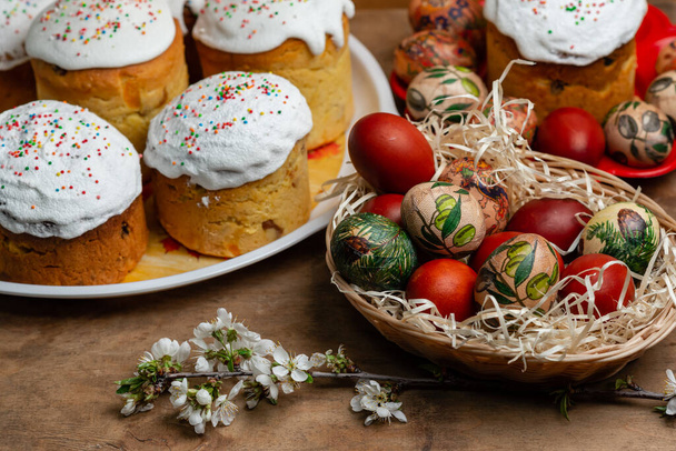 Ciasta wielkanocne i pisanki w wiklinowym koszu z kwitnącą gałązką na drewnianym tle - Zdjęcie, obraz