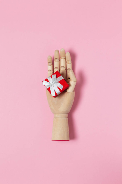 Wooden hand holds small red gift box with white bow on pastel pink background, copy space, flat lay. March 8, February 14, birthday, Valentine's, Mother's, Women's day celebration concept. - Foto, imagen