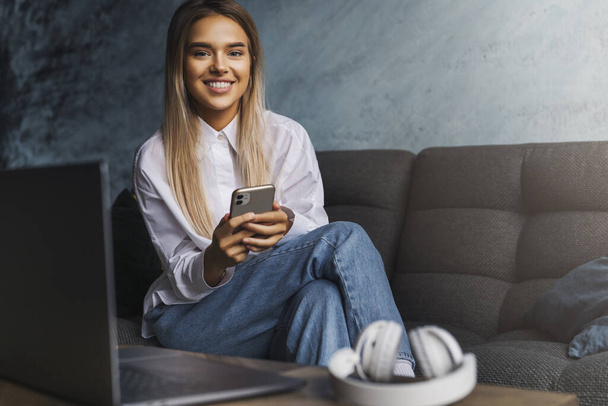 Happy smiling girl sitting on sofa, holding smartphone in hand. Young woman installing messenger app to communicate - Valokuva, kuva