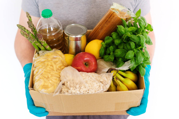Tıbbi kauçuk eldivenli bir erkek, içinde ürünler olan bir karton kutu tutuyor. Ayçiçeği yağı, konserve yiyecek, makarna, yulaf ezmesi, pirinç, sebze ve meyve. Gıda Teslimatı, Yiyecek Bağışı - Fotoğraf, Görsel