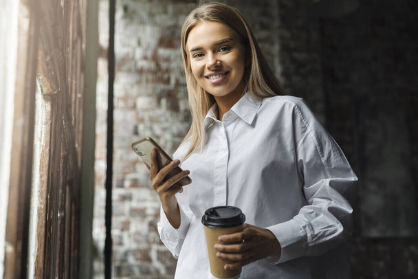 Smiling casually dressed girl holds smartphone and cup of coffee in her hands. Girls use mobile phone for work - 写真・画像
