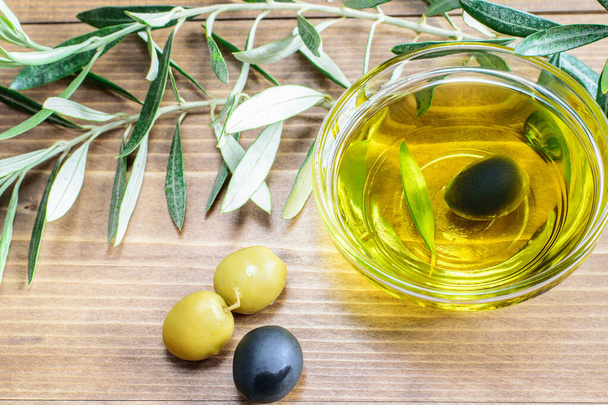 Černá oliva ve skleněné průhledné misce. Některé olivy a olivovníky větve na dřevěném pozadí ve slunečním světle. Koncept zdravé stravy.  - Fotografie, Obrázek