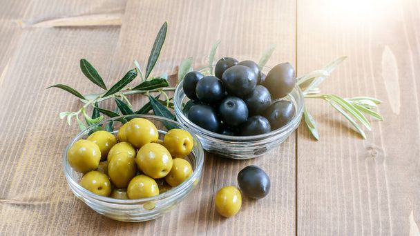 Black and green olives in glass transparent bowl, olive tree branches on the wooden table in sunlights. Healthy diet food concept.  - Photo, Image