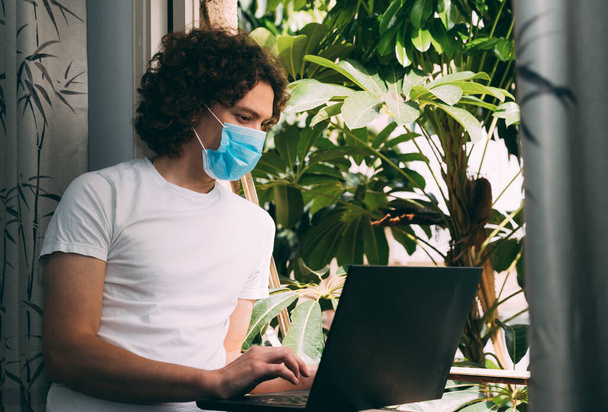 Jonge krullende man met een medisch masker en een wit T-shirt bestelt online eten via een laptop. Hij staat bij een raam van waaruit groene bomen zichtbaar zijn. Blijf thuis. Grijze gordijnen. Kopieerruimte - Foto, afbeelding