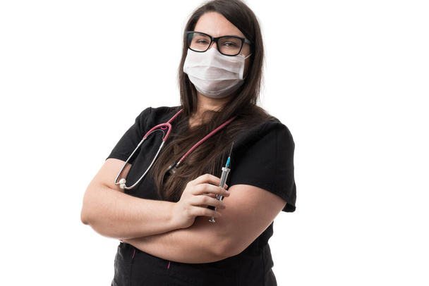 Doctora joven en uniforme negro con una máscara, sosteniendo una jeringa de metal retro, aislada sobre fondo blanco
 - Foto, Imagen
