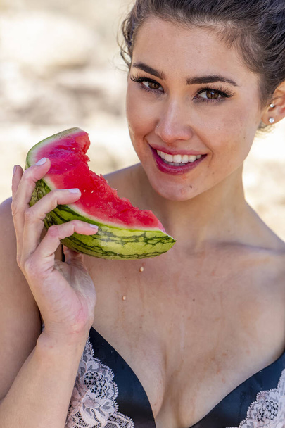Een prachtige brunette bikini model genieten van watermeloen op het strand op een zonnige dag - Foto, afbeelding
