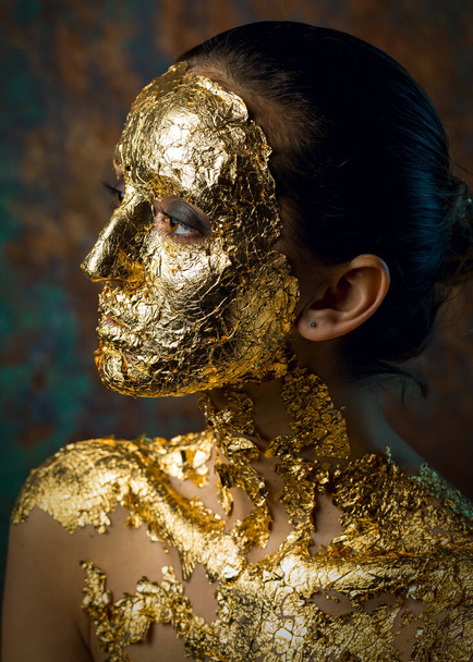 Girl with a mask on her face made of gold leaf. Gloomy studio portrait of a brunette on an abstract background. - Foto, Imagen