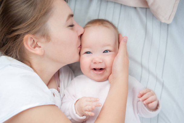 Mother kissing her infant baby in bed. Happy mother kisses baby lying on the bed. Beautiful smiling girl lying on her back. Top view. Flat Lay - Foto, Bild