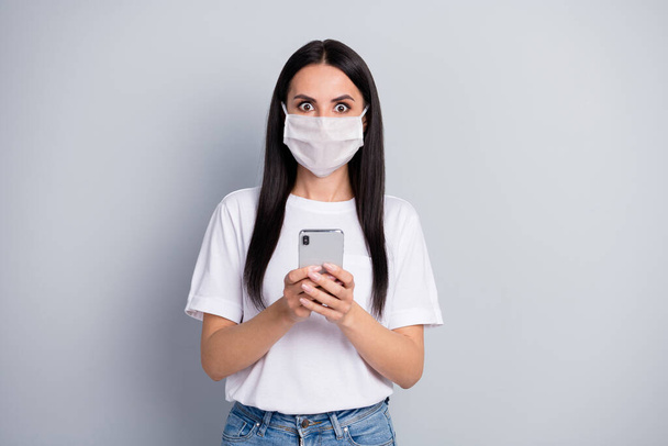 Astonished worried panic girl in respiratory mask use cellphone read social network news impressed corona virus pandemic infection wear t-shirt denim jeans isolated gray color background - Foto, afbeelding