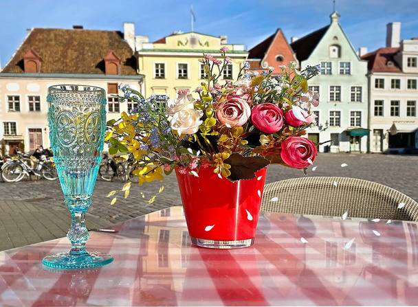 ボトルシャンパンブルーのガラス花バラテーブル上の赤い花瓶ストリートカフェタリン旧市庁舎広場エストニア春の街パノラマ鳥が青空中世都市の夏を飛ぶ  - 写真・画像
