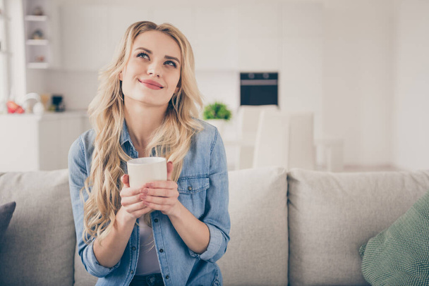 Photo of domestic pretty cheerful blond lady relaxing sit comfy couch holding hot beverage mug dreamer look up enjoy quarantine stay home good mood living room indoors - Фото, зображення