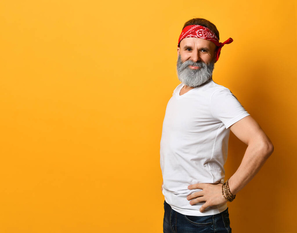Aged male in red bandana, white t-shirt and bracelet. He smiling, put hands on hips, posing sideways against orange background - Фото, изображение