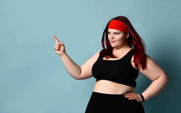 Overweight redhead woman in red headband, black top and leggings. She pointing at something, posing on blue studio background - Foto, afbeelding