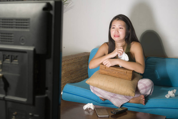 funny lifestyle portrait of young cute and emotional Asian Chinese woman watching korean drama show on TV sitting at home living room couch crying wiping tears with tissue paper - Foto, imagen