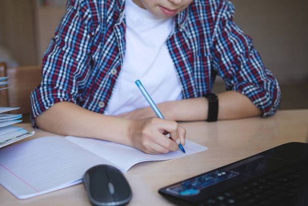Teini-ikäinen poika pöydässä kannettavan tietokoneen ajatus ja kirjoittaa kynällä. Koronavirus on karanteenissa. Valikoiva keskittyminen. Sumea tausta
. - Valokuva, kuva