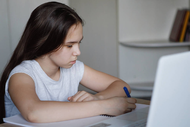 ノートパソコン付きのテーブルの十代の女の子はノートブックにペンで書き込みます。コロナウイルスは家庭教育を隔離した。選択的フォーカス。ぼかしの背景. - 写真・画像