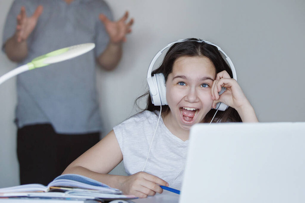 Chicas adolescentes riendo en auriculares en una mesa con un ordenador portátil. Padre borroso agitando sus manos en el fondo. El Coronavirus puso en cuarentena la educación en el hogar. Enfoque selectivo. Fondo desenfocado
. - Foto, imagen