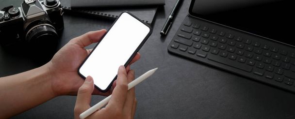 Cropped shot of male entrepreneur using mock-up smartphone while working with digital tablet, camera and other supplies on black table  - Photo, Image