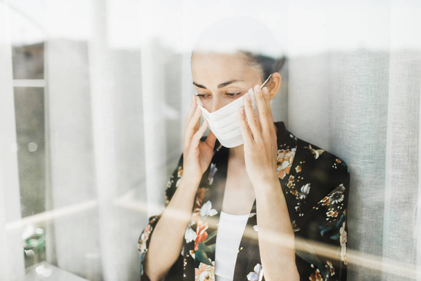 Self isolation in coronavirus quarantine. Woman wearing medical mask. View through window. Prevention of virus spread. - Foto, afbeelding
