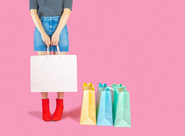The female body part wore denim skirt and red boots. Carrying a pink shopping bag and many pastel colors on pink background selective focus,shopping concept - Fotoğraf, Görsel