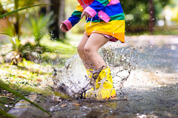Dzieciak bawiący się w deszczu w jesiennym parku. Dziecko skaczące w błotnistej kałuży w deszczowy jesienny dzień. Mała dziewczynka w butach przeciwdeszczowych i tęczowej kurtce na zewnątrz pod ciężkim prysznicem. Obuwie i płaszcz dla dzieci wodoodporne. - Zdjęcie, obraz
