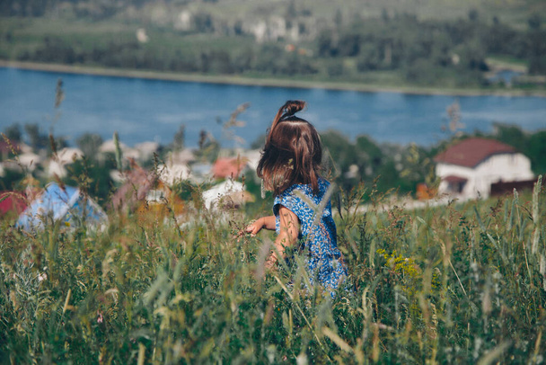 a little girl in a loose dress with loose hair walks in a meadow. Village houses, forest and river as background. The concept of summer, warmth, freedom, village life, sunburn, chldhood - Photo, Image