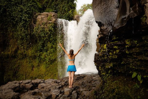 Giovane viaggiatrice in costume da bagno alla cascata. Donna caucasica eccitata che alza le braccia davanti alla cascata. Vista da dietro. Cascata di Tegenungan. Concetto di viaggio. Ubud, Bali, Indonesia
 - Foto, immagini