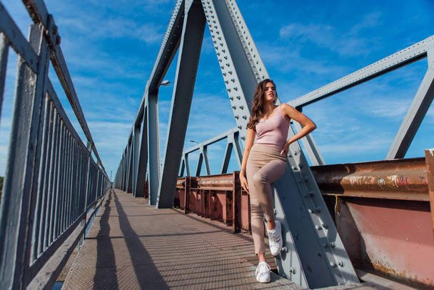 Warm summer portrait of a young beautiful woman posing on the old rusty transport bridge over the river during sunset. - Zdjęcie, obraz