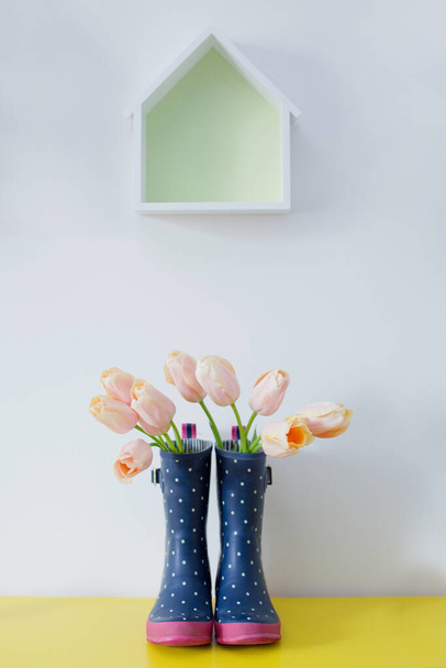 Букет рожевих тюльпанів у блакитних гумових чоботях і порожня полиця у формі будинку на білому тлі
 - Фото, зображення