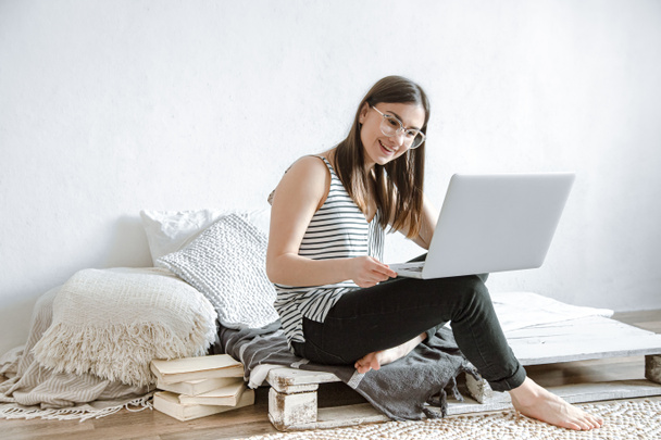 Μια νεαρή γυναίκα δουλεύει εξ αποστάσεως σε έναν υπολογιστή στο σπίτι. Ελεύθερος επαγγελματίας και εργασία στο Διαδίκτυο. - Φωτογραφία, εικόνα