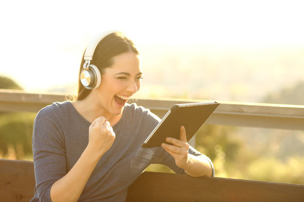 Garota excitada com fones de ouvido e tablet celebrando notícias ao pôr do sol em um parque
 - Foto, Imagem