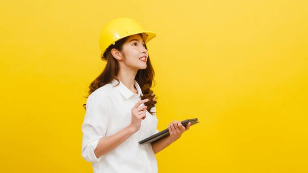 Jonge Aziatische vrouw ingenieur met behulp van digitale tablet met positieve uitdrukking, gekleed in casual kleding en kijken naar camera over gele achtergrond. Gelukkig schattig blij vrouw verheugt succes. - Foto, afbeelding