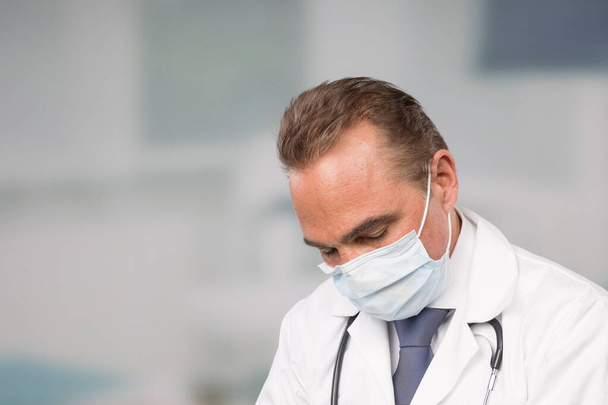 sobre el médico masculino tenso con bata de laboratorio y corbata frente a una sala de la clínica
 - Foto, Imagen