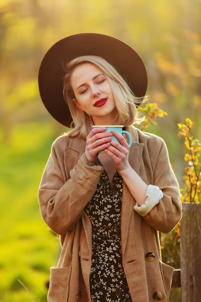 Styl blondýny žena v klobouku a bunda s šálkem kávy v zahradě v západu slunce - Fotografie, Obrázek