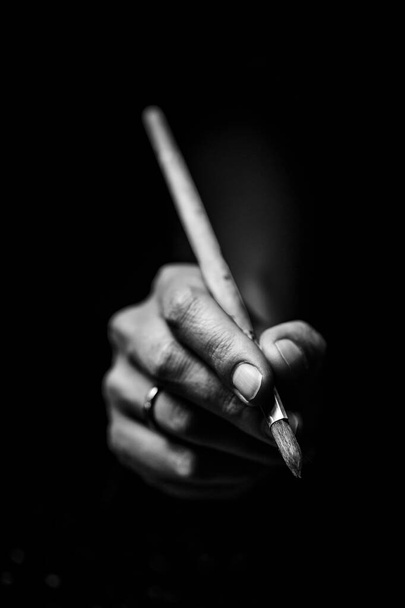 The artist's hands hold a brush. On a black background with deep shadows and bright highlights. Black and white conceptual image - Photo, Image