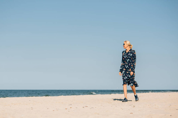 Fin de la cuarentena. Mujer rubia joven en gafas de sol y vestido largo caminando en la playa
  - Foto, Imagen