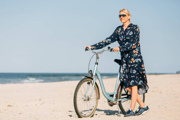 End of quarantine. Young blonde woman in sunglasses and long dress walking with bicycle on beach  - 写真・画像